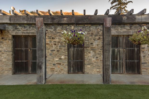 Exterior view of a rustic stone outbuilding or courtyard structure with weathered timber posts, heavy timber beams, dark vertical-plank doors, and exposed roof rafters. Hanging flower baskets add colour above a simple concrete walkway, with a neatly trimmed lawn in the foreground.