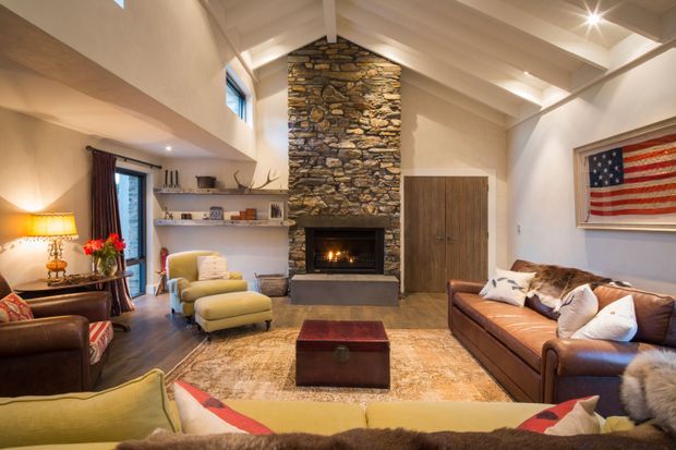 Warm, spacious living room with a vaulted exposed-beam ceiling, prominent natural stone fireplace surround, built-in open shelving, timber cabinetry, leather sofas and lounge chairs, and layered area rugs. A large American flag artwork adds a distinctive accent, while recessed and pendant-style lighting create a cozy ambience.