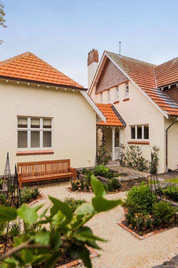 A landscaped courtyard garden enclosed by a pale rendered, multi-wing residential building. The building features steeply pitched tiled roofs in terracotta and grey tones, gabled forms, exposed brick chimney elements, timber-framed windows, and a small covered entry porch. Gravel and paved garden paths surround raised planting beds, with a timber bench and low stone retaining walls.