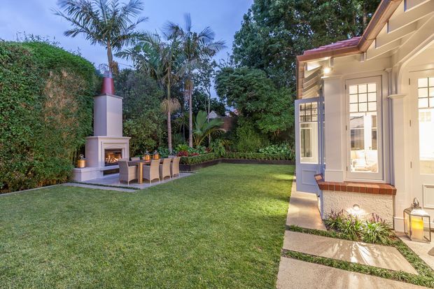 A landscaped backyard viewed at dusk, featuring a manicured lawn, tropical planting and tall hedges, a built-in outdoor fireplace with a chimney and dining table, and the cream-painted exterior of a traditional-style house with large multi-pane windows and a small covered entry.