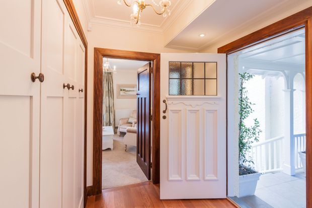 A refined traditional entry hall viewed from inside, featuring white paneled built-in cabinetry, warm timber flooring and dark timber door and trim surrounds. The ornate white front door has decorative raised panels and textured glass, opening toward a covered veranda. A sitting room is visible through the adjacent doorway, and the hall includes decorative ceiling mouldings and a small chandelier.