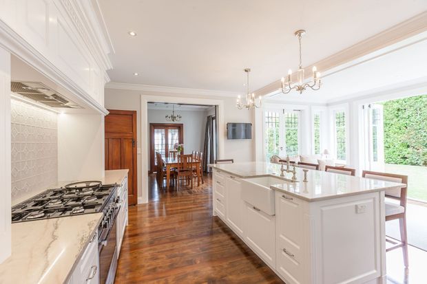 Bright, spacious traditional kitchen with white Shaker-style cabinetry, a large central island, marble-look benchtops, patterned tiled splashback, stainless steel range cooker, and polished timber flooring. The kitchen opens to a dining area and a light-filled glazed breakfast or sunroom with garden views. Decorative chandeliers, ornate ceiling mouldings, and detailed cabinetry create a refined classic character.