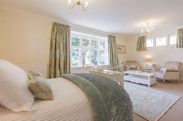 Bright, traditional bedroom with a large bed in the foreground, pale quilted bedding, and muted sage-green throw and cushions. The room features elegant bay-style multi-pane windows with patterned curtains, high decorative cornices, chandeliers, a seating area with upholstered armchairs, a white carved coffee table, and a soft area rug. The overall atmosphere is refined, calm, and light-filled.