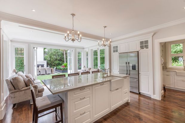 Bright, refined traditional kitchen with a large white island, integrated seating, paneled cabinetry, stainless-steel refrigerator, polished stone benchtop, hardwood flooring, and classic chandelier lighting. French doors open to a landscaped outdoor area, while a butler-style pantry or utility space is visible through the doorway on the right.