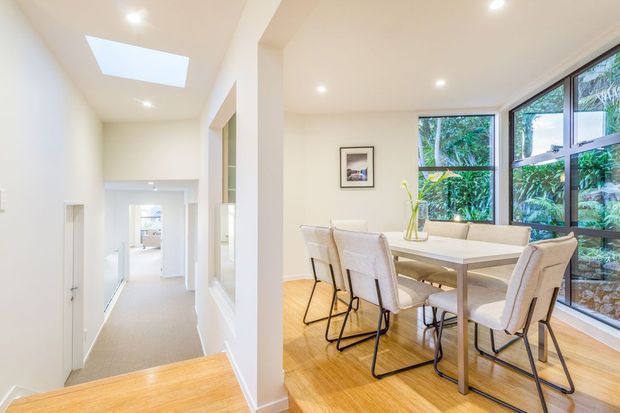 Bright, contemporary dining area opening onto a white hallway, with a light timber floor, white walls, recessed lighting, skylight, and large black-framed windows overlooking lush greenery. A simple rectangular dining table with upholstered chairs occupies the main space.