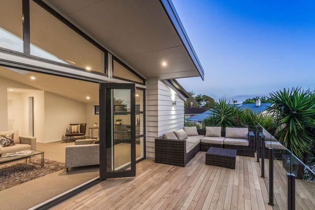 Contemporary residential terrace with expansive timber decking, glass balustrade, and an outdoor lounge furnished with dark woven seating. The open corner glazing and black-framed doors connect the deck to a bright interior living room, while horizontal exterior cladding and a broad roof overhang define the modern facade. Tropical-style planting and a distant coastal-looking view frame the outdoor area.