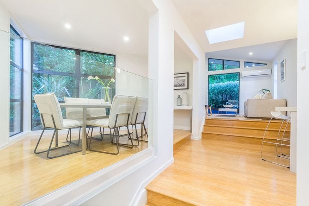 Bright open-plan interior with a timber staircase and polished light timber flooring, opening onto a dining area enclosed by a glass balustrade. The dining space has a white table and contemporary white chairs beside large windows overlooking greenery. A raised living or sitting area is visible beyond, with built-in shelving, a skylight, and air conditioning.