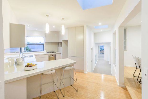 Bright contemporary open-plan kitchen with a large white island and integrated seating, pale built-in cabinetry, timber flooring, pendant lights, skylights, and adjoining circulation spaces. A dining area is visible through the opening on the right.