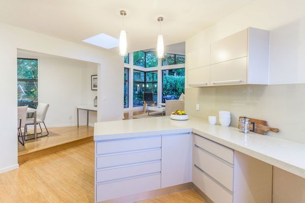 Bright open-plan kitchen with a large L-shaped island and peninsula, white flat-panel cabinetry, pale stone benchtops, timber flooring, pendant lights, skylight, and extensive corner glazing overlooking greenery. A dining area and living space are visible beyond the kitchen.