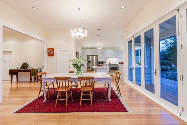 Bright open-plan dining area with a long light-wood dining table, spindle-back wooden chairs, and a white tablecloth positioned over a patterned red rug. The room has pale cream walls, polished timber flooring, white traditional-style trim and cabinetry, recessed ceiling lights, a crystal chandelier, and large white-framed glazed folding doors opening to a timber deck. A compact white kitchen is visible beyond the dining space.