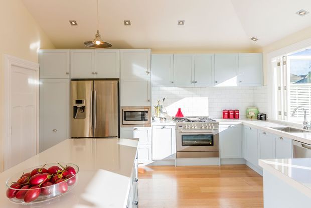 Bright, spacious white kitchen with extensive shaker-style cabinetry, stainless-steel appliances, white subway-tile splashback, pale timber flooring, and white stone benchtops. A central island and peninsula create generous preparation and dining space, with natural light entering through the window and glazed door. The room has a clean, welcoming, traditional-meets-contemporary character.
