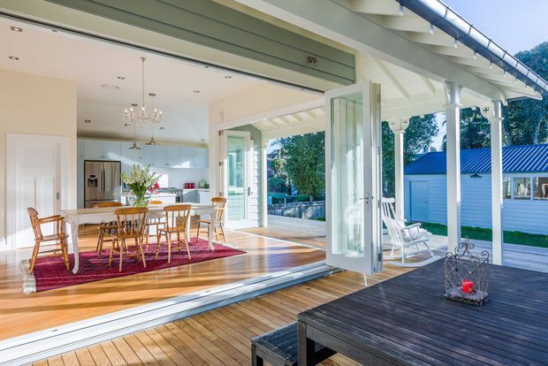 Bright coastal-style open-plan dining and kitchen interior opening onto a timber deck through fully glazed folding or stacking doors. The room features white walls and cabinetry, exposed painted timber veranda structure, polished timber flooring, a timber dining table with spindle-back chairs, pendant lighting, and a red patterned rug. The deck overlooks a landscaped outdoor area and a small blue-roofed outbuilding.