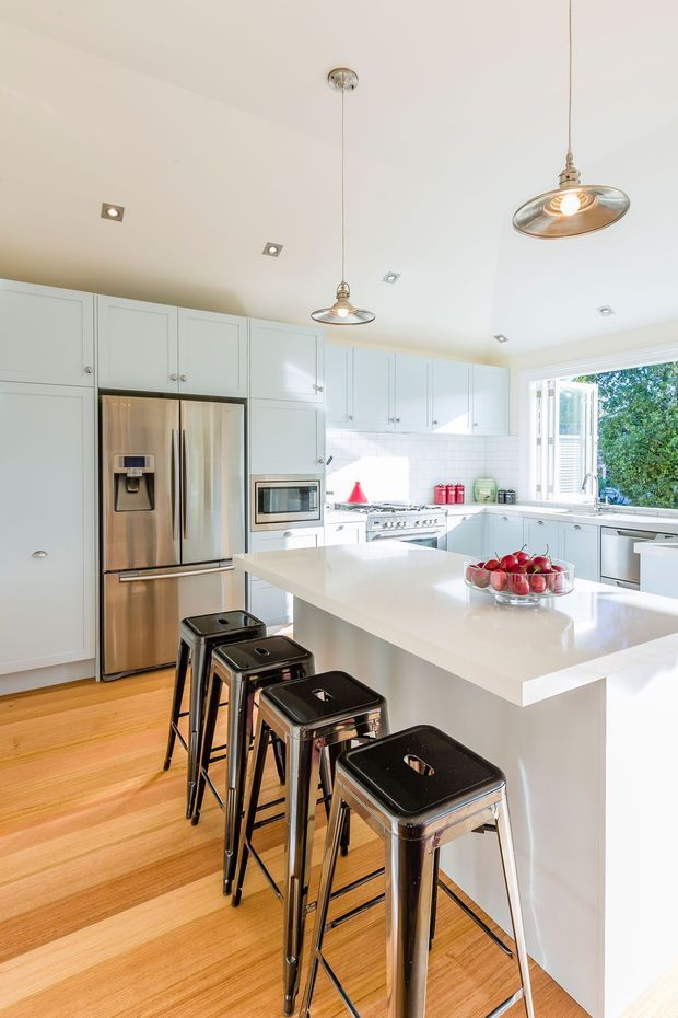 Bright, spacious white kitchen with extensive shaker-style cabinetry, stainless-steel appliances, a large white island with seating for four or more, and light timber flooring. A white subway-tile splashback, recessed ceiling lights, and polished metal pendant lights contribute to the clean, contemporary look. Large windows bring in abundant daylight and provide views of greenery.