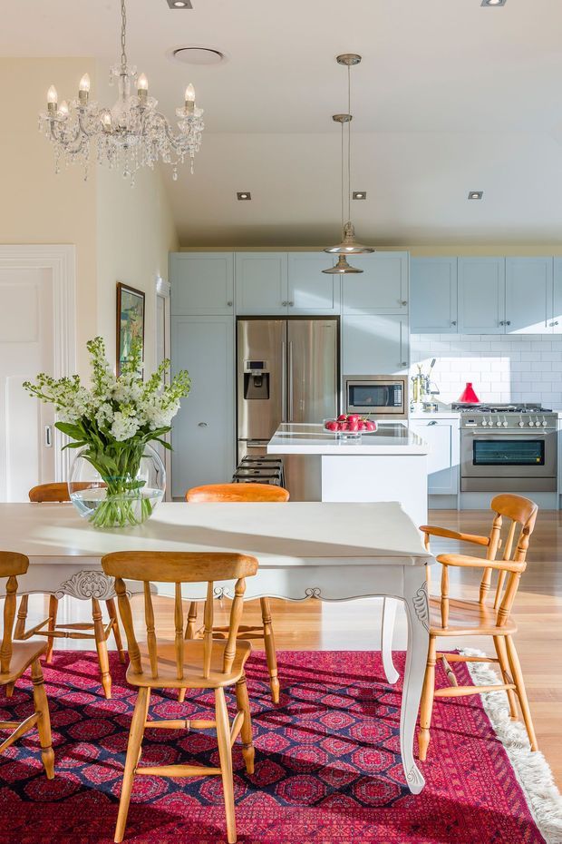Bright, elegant kitchen and dining area with pale blue built-in cabinetry, stainless-steel appliances, white subway-tile splashback, a central island, and a large white carved-leg dining table. Warm timber flooring, traditional wooden chairs, a crystal chandelier, pendant lights, and a richly patterned red Persian-style rug create a refined yet inviting interior.
