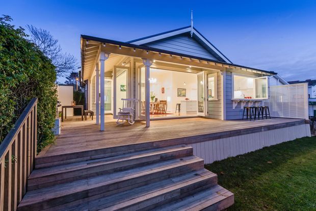 Twilight exterior view of a light-clad, single-storey gable-roofed home with a broad timber deck and covered veranda. Large glazed openings reveal a bright open-plan kitchen and dining area, while the deck includes steps, seating, and a built-in outdoor bar or kitchen counter.