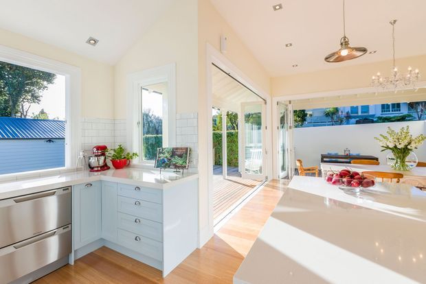 Bright open-plan kitchen and dining area with white cabinetry, white stone benchtops, stainless-steel appliances, pale timber flooring, and white subway-tile splashback. Large glazed doors open to a timber deck, bringing in abundant natural light. The dining area includes a timber table and chandelier, while the kitchen features pendant lighting and a central island or peninsula.
