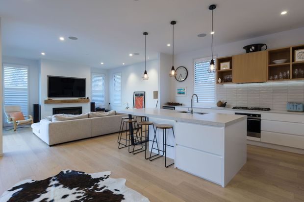 Open-plan contemporary kitchen and living area with a large white island, integrated seating, white cabinetry, textured tile splashback, pendant lighting, and timber-look flooring. The living zone includes a sectional sofa, wall-mounted television, fireplace, and cowhide rug, with abundant windows and soft natural light.