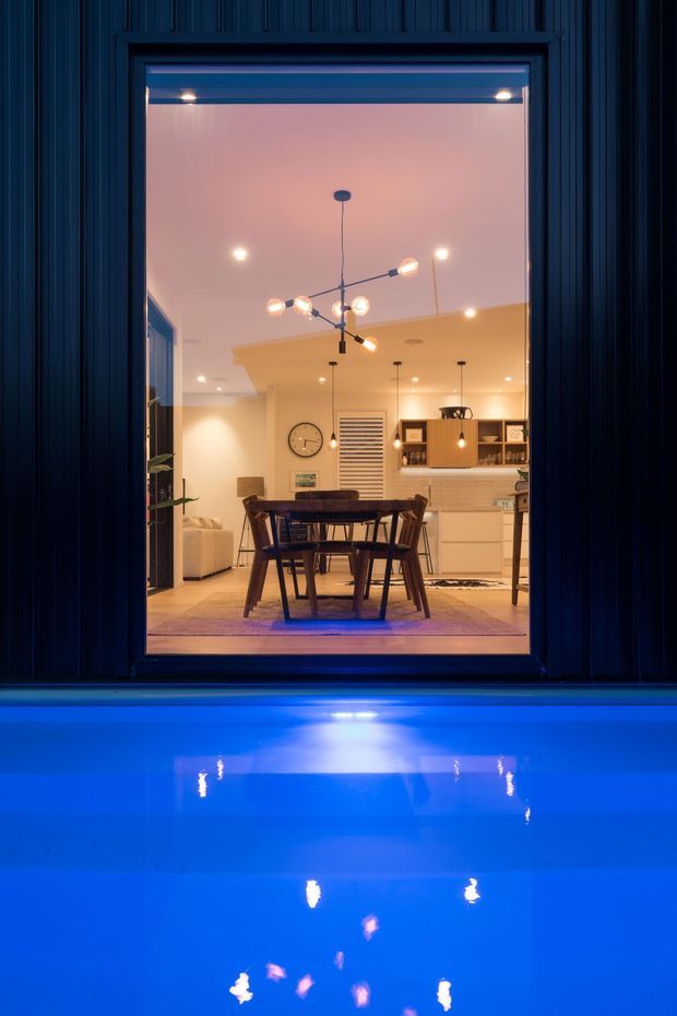 Nighttime view toward a warmly lit dining and kitchen area through a large framed opening, with a dark vertically clad exterior surround in the foreground and vivid blue pool water reflecting the interior lights below. The interior features a timber dining table and chairs, pendant and sculptural ceiling lighting, pale cabinetry, open shelving, and a light neutral palette.