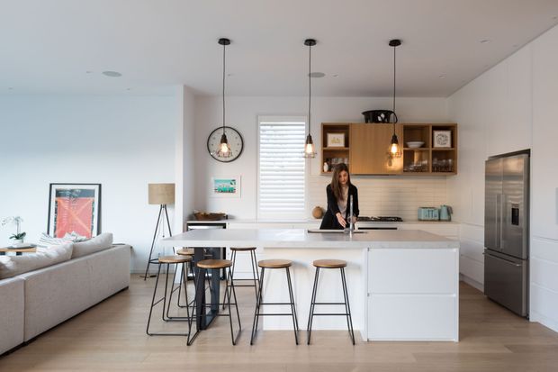Bright open-plan kitchen with a large white island and seating for four, integrated timber cabinetry, stainless-steel refrigerator, pendant lighting, and pale timber flooring. A living area is visible to the left, with clean contemporary detailing and abundant natural light.
