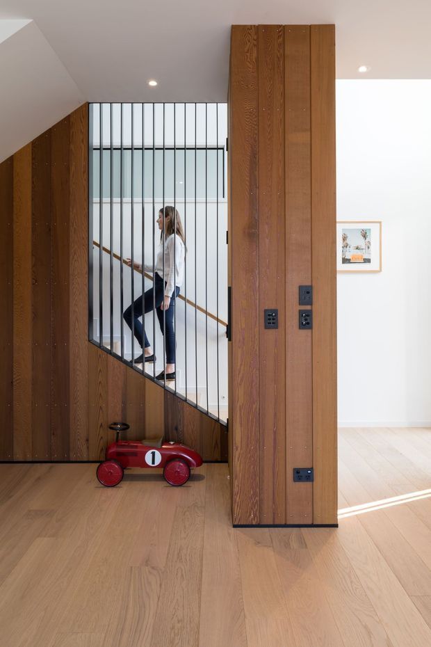 A bright contemporary residential hallway and stairway featuring extensive vertical timber wall panelling and a matching central structural partition. A black metal balustrade encloses the open stair, with pale timber flooring, white walls and ceiling, recessed lighting, and a framed artwork visible in the adjacent room. A small red toy car sits beneath the stairs.