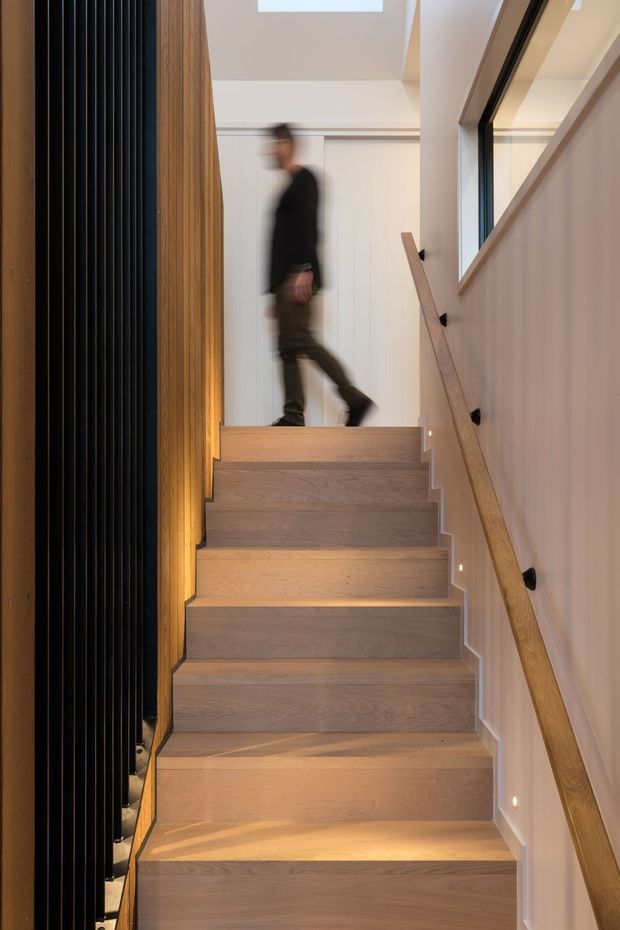 A contemporary interior stairway with warm light-oak timber treads and matching handrail, white walls, integrated step lighting, a tall black vertical slatted feature wall on the left, and a black-framed internal window on the right. A blurred person is walking across the upper landing.