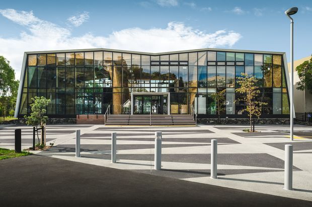 A contemporary low-rise commercial building with a broad glazed facade, dark structural framing, and a gently folded roofline. A central recessed entrance is reached by wide steps, opening onto a patterned paved forecourt with bollards and young trees. Interior spaces and structural elements are visible through the extensive glazing.