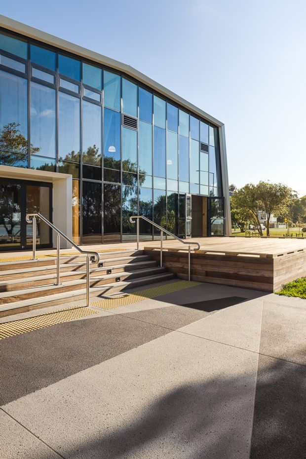 Exterior view of a contemporary public or commercial building with a tall, curved glazed facade, dark metal framing, and a shallow projecting roof edge. Broad steps and an accessible ramp with stainless-steel handrails lead to the entrance. A raised timber-clad platform and exposed aggregate or concrete paving occupy the forecourt.