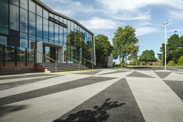Exterior view of a contemporary institutional or commercial building with a sweeping glazed curtain wall, dark structural framing, broad entrance steps and accessible handrails. The building fronts a large geometric paved forecourt with alternating light and dark triangular bands, surrounded by mature trees and open landscaped grounds.