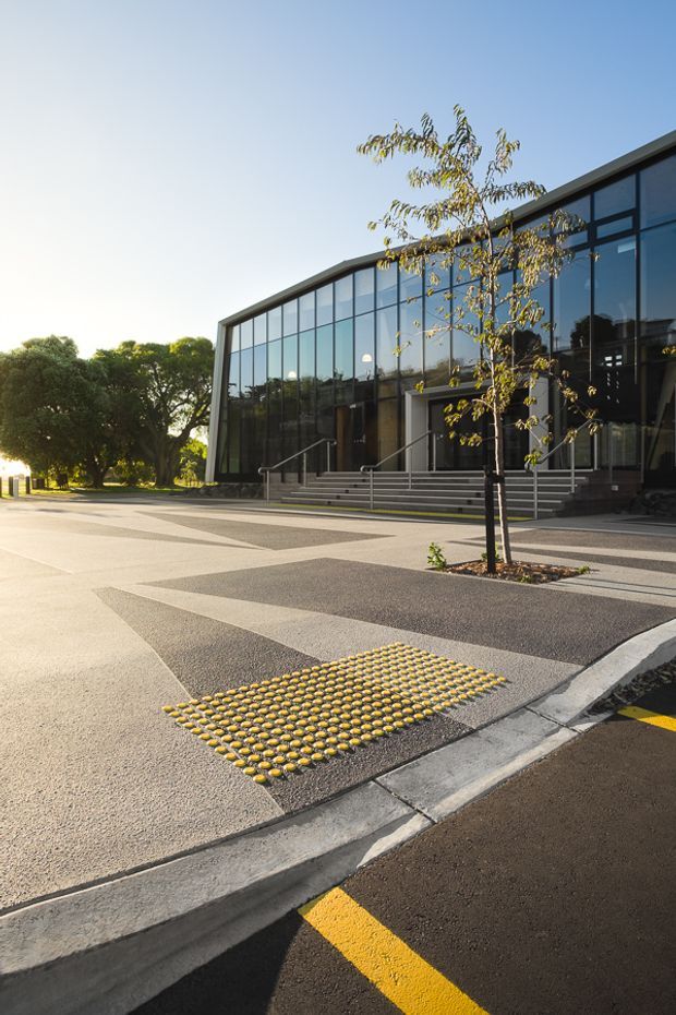 Exterior view of a contemporary glass-fronted institutional or commercial building with broad steps, accessible tactile paving, a landscaped forecourt, and patterned asphalt paving. The low-rise facade features extensive dark-framed glazing and a gently sloping roofline.