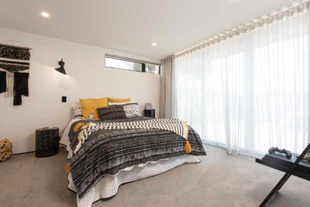 Bright contemporary bedroom with a low-profile bed dressed in layered black, white, mustard-yellow, and patterned textiles. The room has soft grey carpet, white walls and ceiling, a black wall sconce, minimalist bedside furniture, and extensive floor-to-ceiling glazing covered by sheer white curtains.