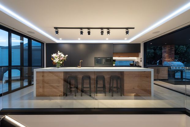 A sophisticated contemporary kitchen centered on a large timber-clad island with integrated seating for four. The space features dark matte cabinetry, a pale stone benchtop, a tiled or polished light floor, extensive black-framed glazing, recessed perimeter LED lighting, track lighting, and a view toward an outdoor entertaining area with barbecue facilities.