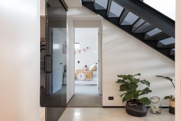Bright contemporary interior hallway beneath a black metal open-riser staircase. The view includes a glass-panel door and partial kitchen cabinetry to the left, a carpeted bedroom visible through the doorway, and potted plants arranged along the stair wall. The space combines white walls, polished light flooring, dark metal stair framing, and warm neutral furnishings.