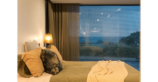 A cozy bedroom viewed toward a large floor-to-ceiling window with horizontal blinds, overlooking a dark outdoor landscape at dusk. The bed is dressed in warm olive-beige bedding with layered pillows, a cream knitted throw, and a floral accent cushion. Full-height curtains frame the glazing, while a small bedside lamp creates a warm, intimate atmosphere.