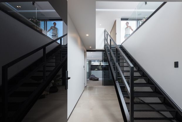 A contemporary interior stairway with two flights of black metal open-tread stairs, dark balustrades, white walls, polished concrete flooring, and extensive internal glazing. A full-height dark reflective wall on the left mirrors the stair and upper landing, while daylight enters from the upper-level windows and glazed doors at the rear.