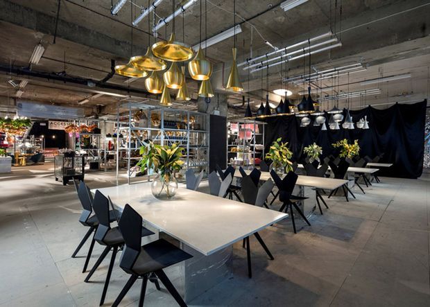 An industrial-style commercial showroom or retail space with exposed concrete ceilings, visible services, and polished concrete flooring. Large communal dining tables with angular black chairs sit beneath clusters of metallic pendant lights, while shelving, plants, and displayed lighting products occupy the background.