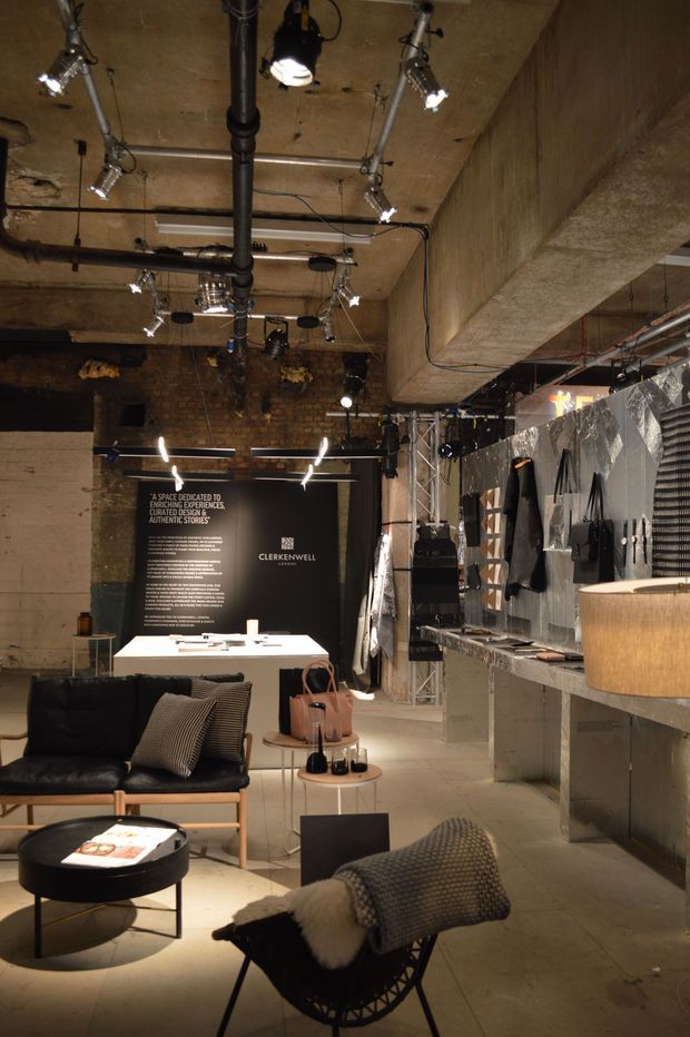 An industrial-style commercial showroom or retail interior with exposed concrete beams and ceiling, visible black piping, exposed brickwork, track and suspended lighting, and raw unfinished surfaces. The space contains a lounge setting with black leather seating, side tables, a desk or display table, shelving and product displays along the right wall, and a large branded graphic panel at the rear.
