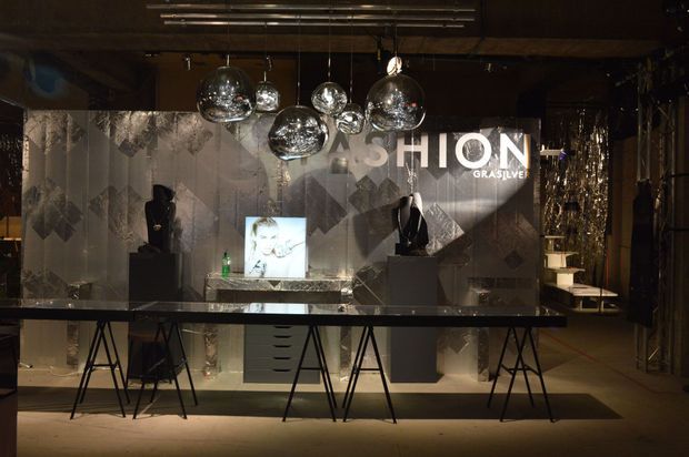 A dark, dramatic fashion retail showroom or commercial display space featuring a long central display counter on trestle-style supports, mannequins or busts on pedestals, illuminated product imagery, reflective geometric wall panels, and clustered chrome pendant globe lights. The space has an industrial-luxe, theatrical merchandising aesthetic.