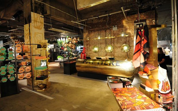 An industrial-style retail or showroom interior with exposed red brick walls, dark structural beams, polished concrete flooring, and warm display lighting. Shelving and tables present colourful merchandise, while a large metallic cylindrical display counter and vibrant hanging textiles create a bold, eclectic focal point.