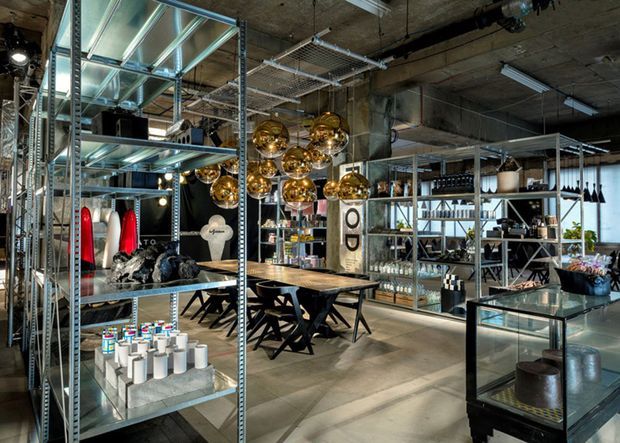 An industrial-style retail showroom or commercial interior with exposed concrete ceilings, metal shelving, glass display cases, and a central dining table beneath clustered brass pendant lights. The space displays homewares, lighting, furniture, and decorative objects in an open, warehouse-like layout.
