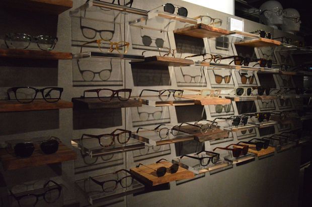 Dimly lit retail display wall showcasing numerous eyeglass frames and sunglasses arranged on floating wooden shelves and wire rack holders. The display has an industrial, handcrafted showroom character with dark shelving and warm accent lighting.