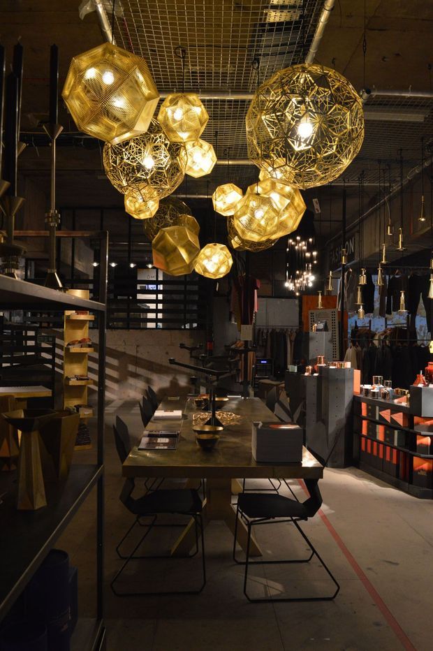 A dimly lit industrial-style commercial interior, likely a showroom or retail space, with a long dining table and black chairs beneath a cluster of ornate gold geometric pendant lights. Exposed ceiling services, metal shelving, dark cabinetry, display counters, concrete flooring, and warm accent lighting create a dramatic warehouse-like atmosphere.
