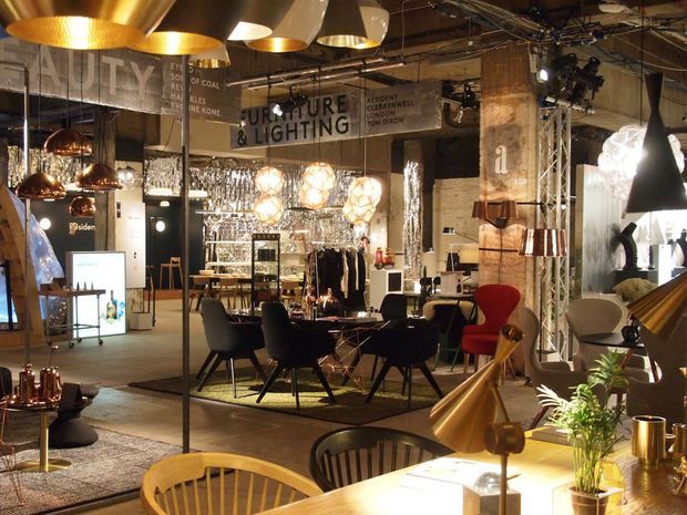 A large, eclectic interior showroom displaying furniture and lighting. The space features exposed ceiling services, industrial truss lighting, concrete or raw structural columns, polished metal and glass shelving, and numerous pendant lights. Dining and lounge furniture is arranged throughout, with warm timber tables, dark upholstered chairs, and a mix of sculptural contemporary furnishings.