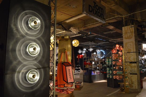 Interior view of an eclectic industrial-style lighting and design showroom, featuring exposed structural elements, rough concrete columns, an unfinished ceiling, metal display shelving, and numerous decorative light fixtures. A large illuminated wall installation dominates the left side, while colourful product displays and hanging pendant lights fill the space.