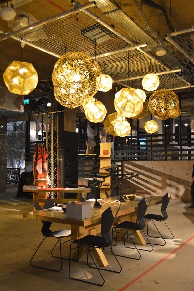 A dramatic industrial-style dining or hospitality interior with exposed services, ductwork, pipes, cable trays, and a raw concrete ceiling. Multiple large sculptural amber pendant lights hang above long timber dining tables surrounded by black angular chairs. The space includes dark timber and slatted partitions, exposed structural elements, and warm directional lighting, creating a bold, atmospheric restaurant or commercial venue.