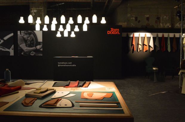 A dimly lit industrial design studio or showroom displaying leather and material samples across a large worktable. A black reception or display wall carries the Tom Dixon logo, while pendant lights hang overhead and additional fabric or material samples are arranged on the right wall. The space has exposed, weathered architectural surfaces and a moody exhibition atmosphere.