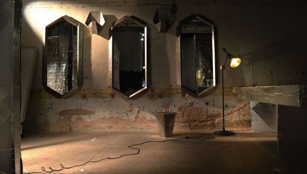 A dim, atmospheric room with distressed exposed brick and peeling plaster walls, polished concrete flooring, three tall faceted wall mirrors, a sculptural central pedestal, and a black adjustable floor lamp. The space has an unfinished industrial gallery or showroom character with warm, dramatic lighting.