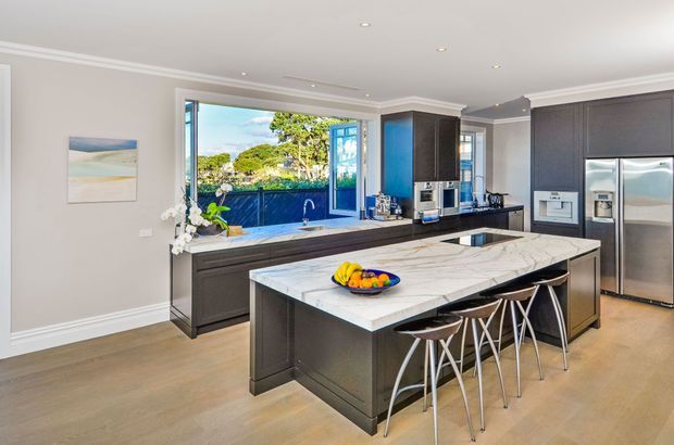 A spacious contemporary kitchen with a large dark island, veined light stone benchtop, integrated appliances, and seating for four. Full-height glazing and an open window connect the room to a leafy outdoor setting, while recessed ceiling lights, light painted walls, and timber flooring create a bright, refined interior.