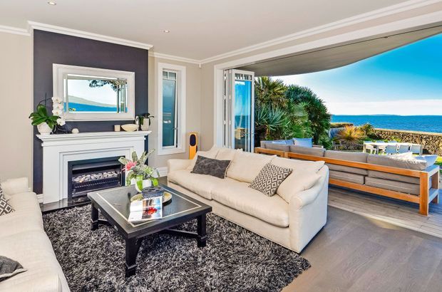 Bright coastal living room with classic white moulded trim, a dark feature wall and fireplace surround, cream sofas, a black coffee table, and a large textured rug. Wide bi-fold doors open directly onto a timber deck with panoramic ocean views and outdoor seating.
