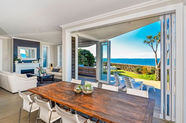 Bright coastal dining and living area opening through wide bi-fold doors to a landscaped outdoor entertaining space with ocean views. The interior features a large rustic timber dining table, white dining chairs, pale upholstered seating, light flooring, white trim, and a fireplace or media feature wall. The outdoor area includes a dining setting and lawn overlooking the sea.