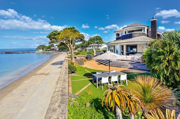 A large multi-level coastal residence directly beside a sandy beach, with dark shingle-like roof sections, light exterior walls, balconies, expansive glazing, and a waterside outdoor entertaining area. The landscaped garden includes a dining table, umbrella, deck, lawn, and sculptural coastal planting. Bright blue sky and calm water create a relaxed beachfront setting.